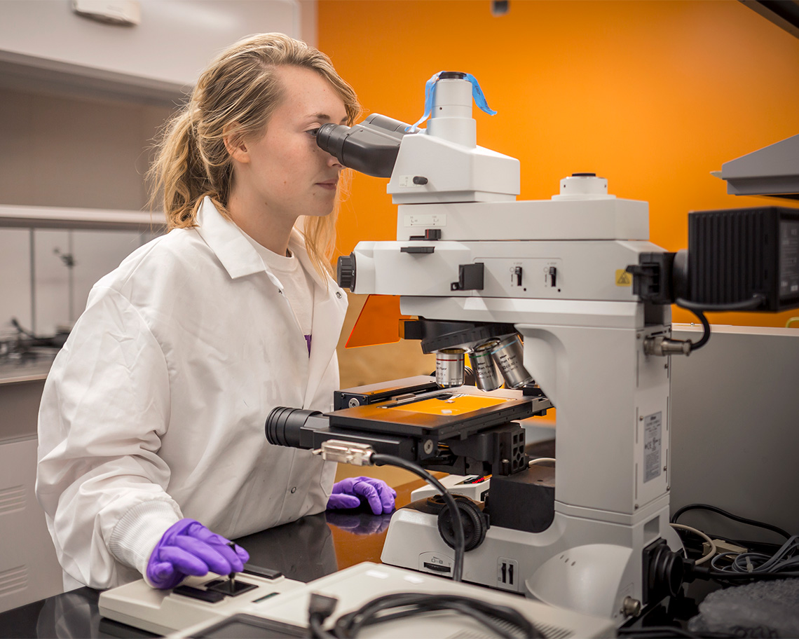 A woman wearing a lab coat looks in a  microscope
