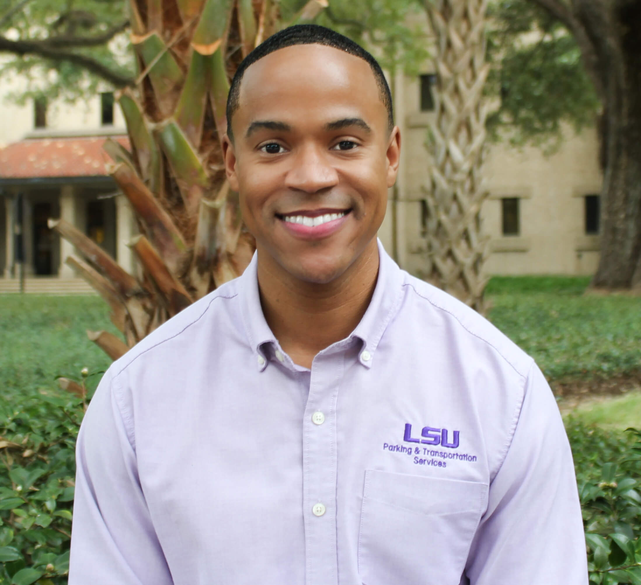 Man in purple shirt smiling and standing in front of a tree and bushes