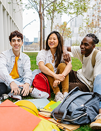 Three students sitting together outdoors with backpacks and school materials.